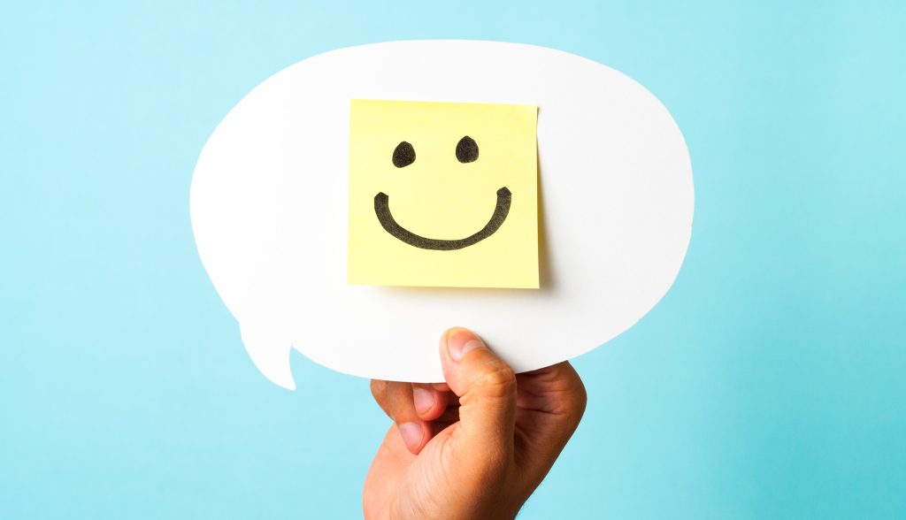 Hand holding white speech bubble with drawing of a happy smiling emoticon on a yellow paper and white background. Happiness, positive feedback photo
